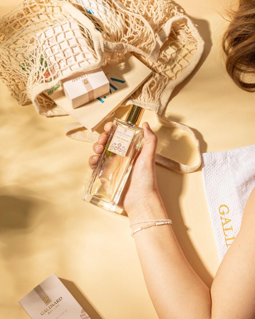 Main tenant un flacon de parfum « L’été dernier » de Galimard posé sur une surface beige avec sac en filet, accessoires d’été et ambiance ensoleillée.